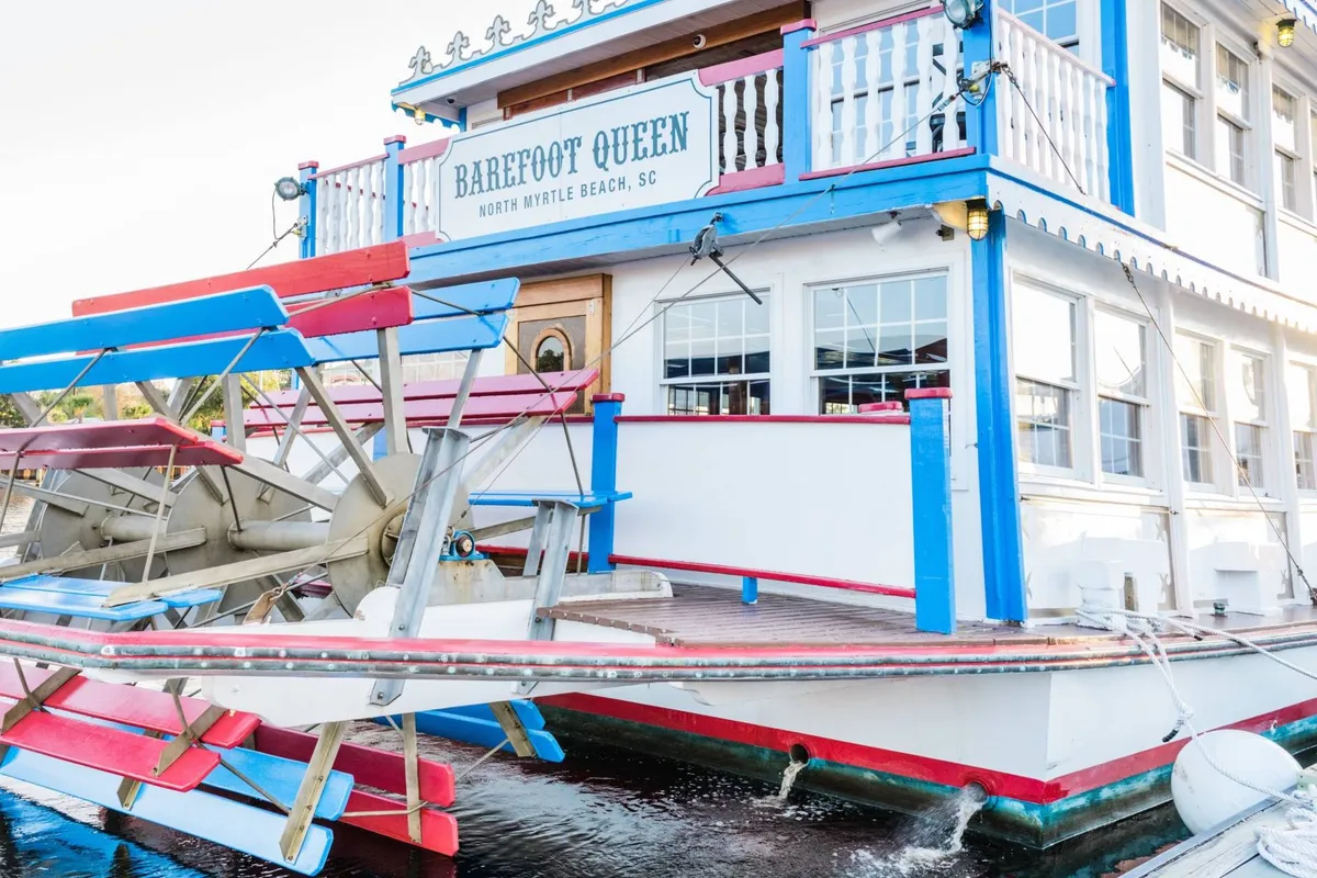 This image shows the colorful and inviting exterior of the 'Barefoot Queen' paddle wheel boat docked at Barefoot Landing, with signage, a vibrant facade, and water in the foreground. The scene suggests a playful, lively atmosphere with possibilities for dining, sightseeing, and live events on the water.
