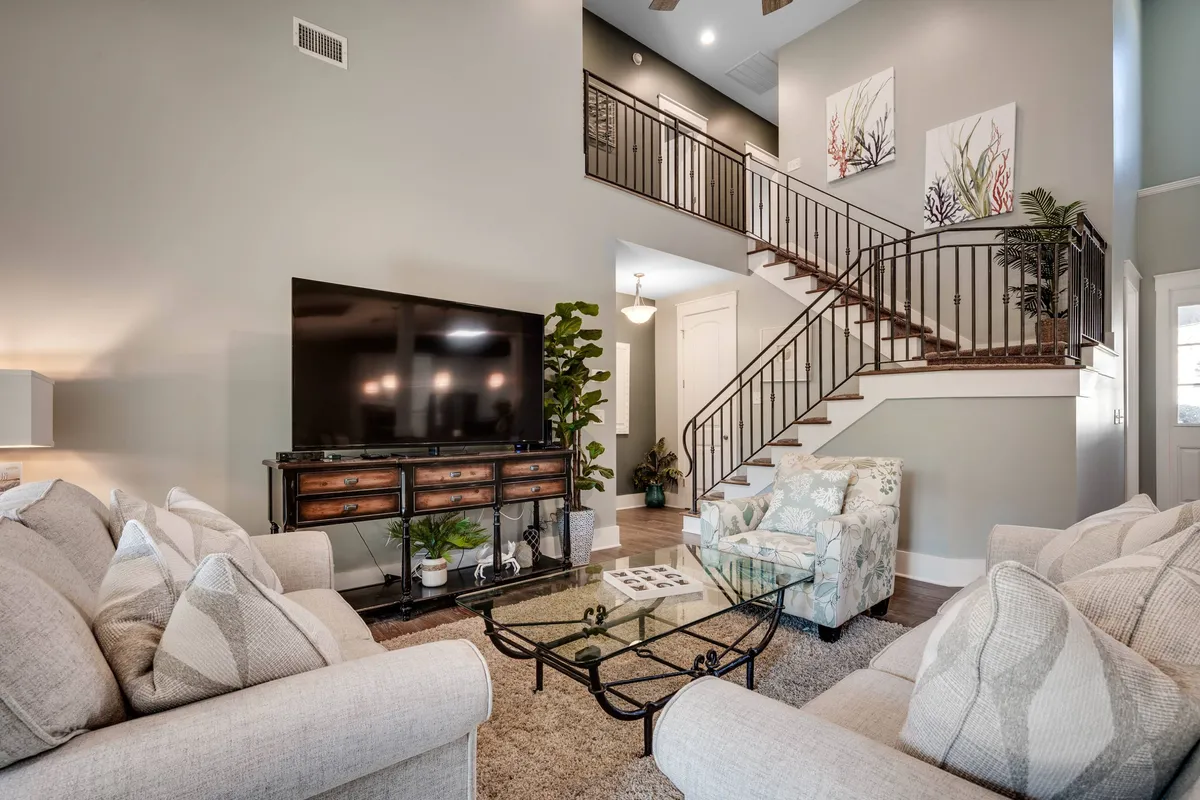 The image showcases a spacious, inviting living room with a comfortable sofa, a stylish coffee table, and a large television. A staircase adds architectural interest, while the cozy layout suggests a perfect spot for relaxing, hosting gatherings, or starting mornings slowly. The decor is modern yet warm, creating a homey, beach-casual vibe.