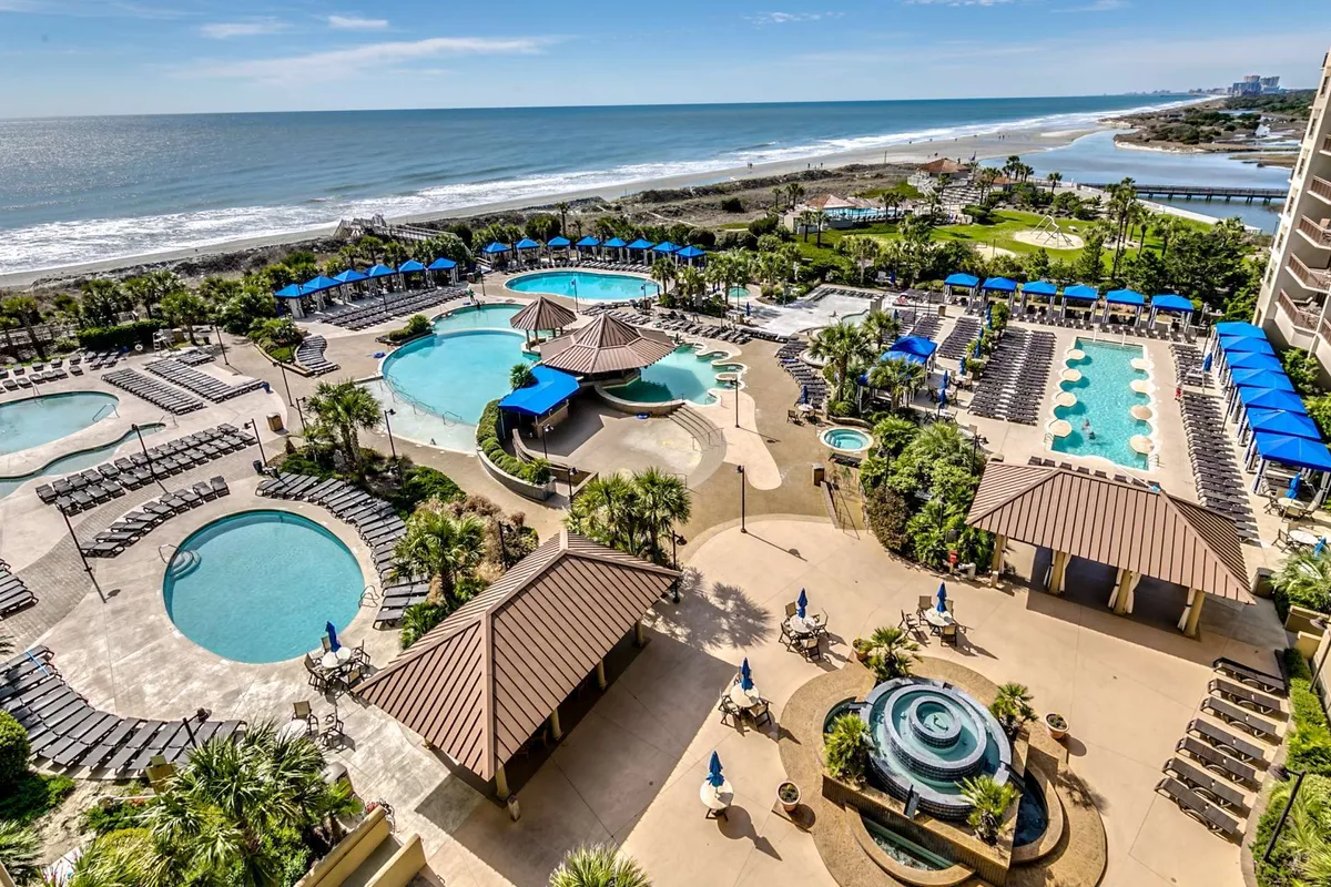 This image features a sprawling resort-style pool complex, complete with multiple swimming pools, beachfront access, palm trees, and a plentiful array of lounge chairs. The scene is bathed in natural light, suggesting a lively yet relaxing atmosphere ideal for sunbathing, swimming, or simply soaking up the tranquil resort environment.