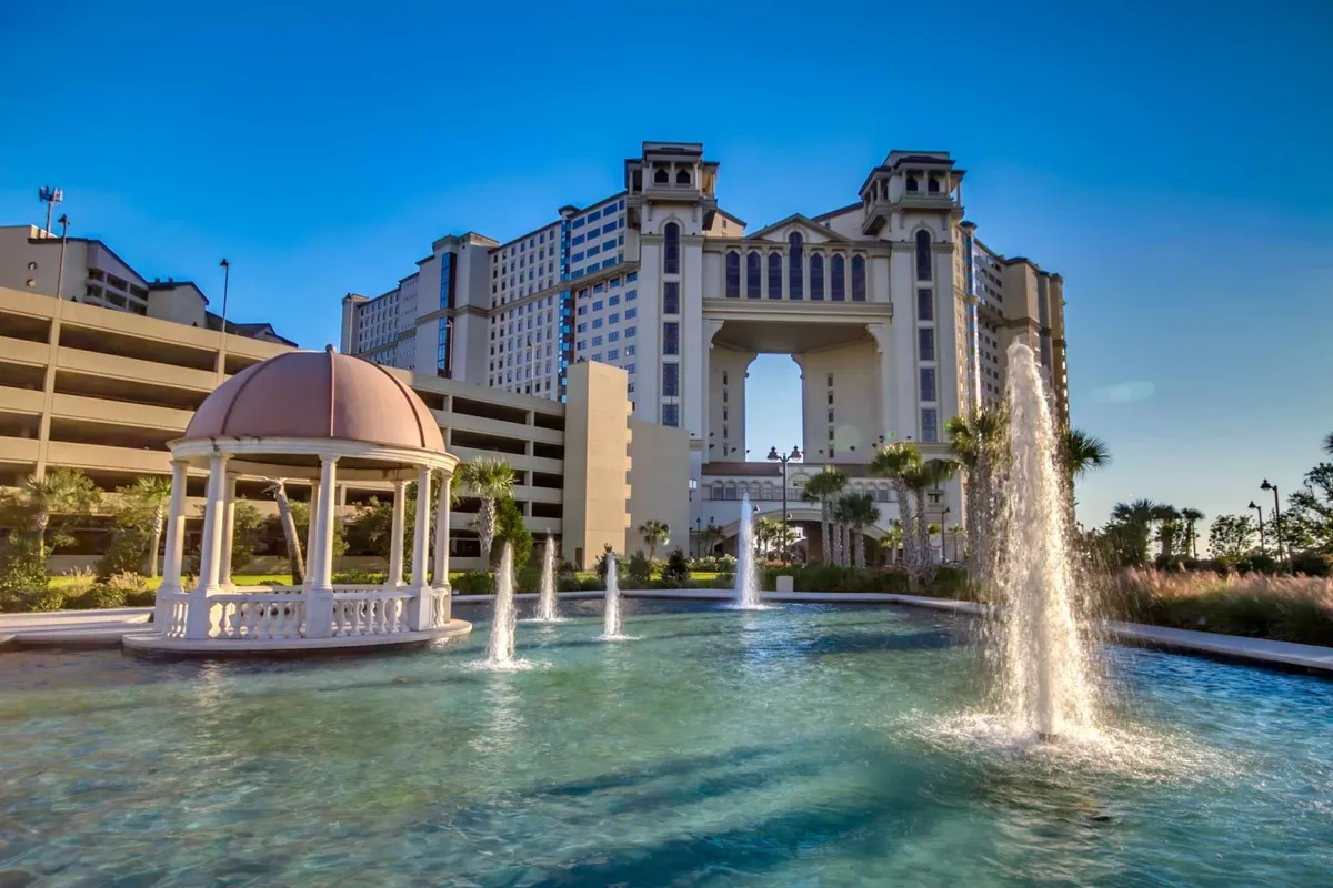 A grand building structure stands behind a decorative gazebo and tranquil fountains, set alongside a serene body of water. The setting evokes a luxurious resort atmosphere, ideal for post-golf relaxation, spa visits, or leisurely strolls near the water.