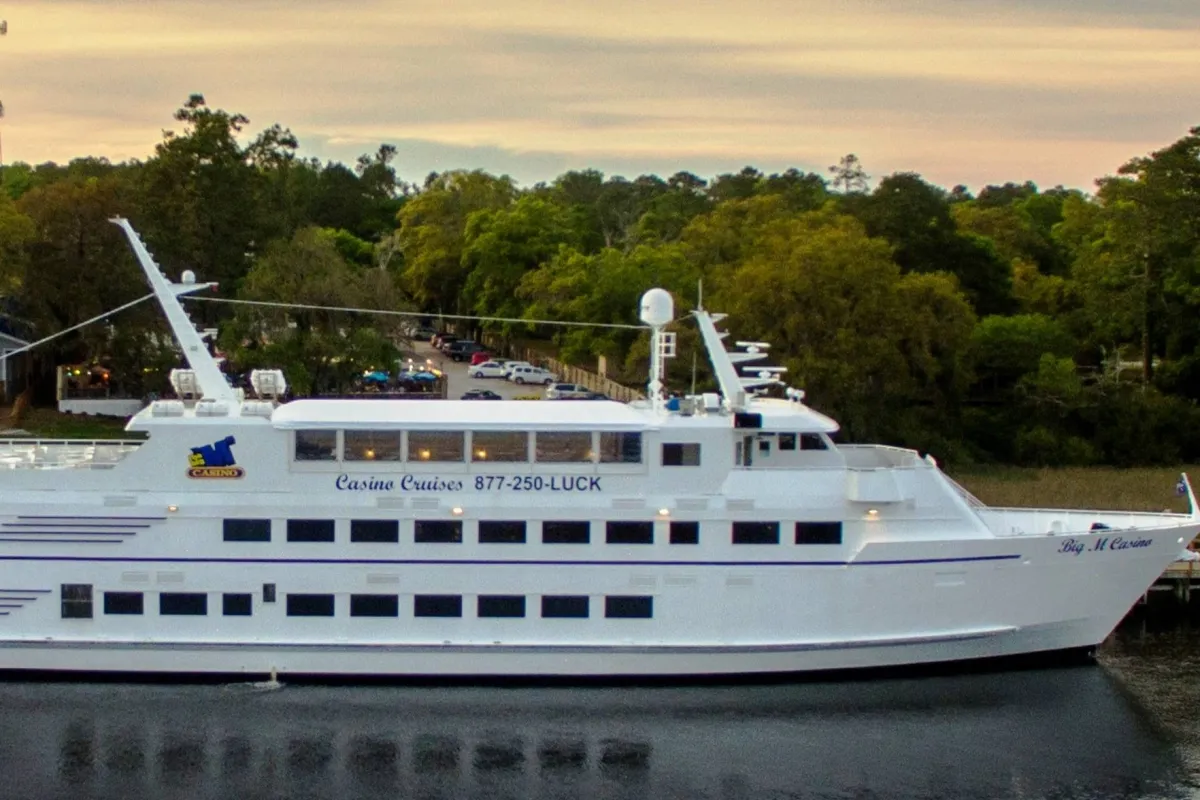 This vibrant exterior shot showcases a large white cruise ship, surrounded by lush greenery and glistening water. Prominent signage indicates a casino and entertainment venue, suggesting opportunities for cruising, dining, and enjoying live events against a scenic sunset backdrop.