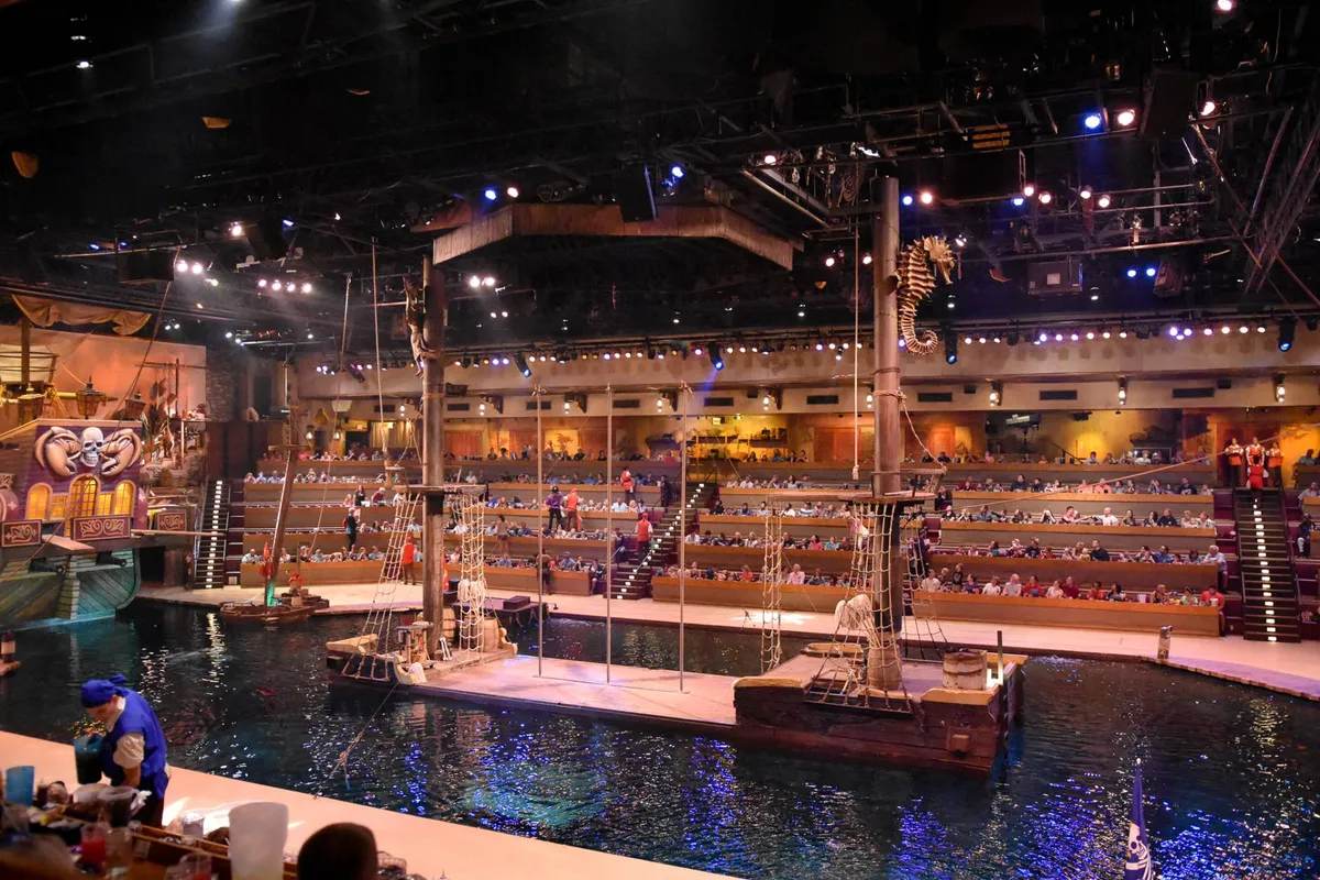 This image captures the excitement of a live performance at the Pirate’s Voyage Show. The scene features a grand pirate ship set on a water-filled arena, surrounded by themed decorations and audience seating. The lively atmosphere is palpable, with guests immersed in the spectacle and the anticipation of audience participation and dining experiences.
