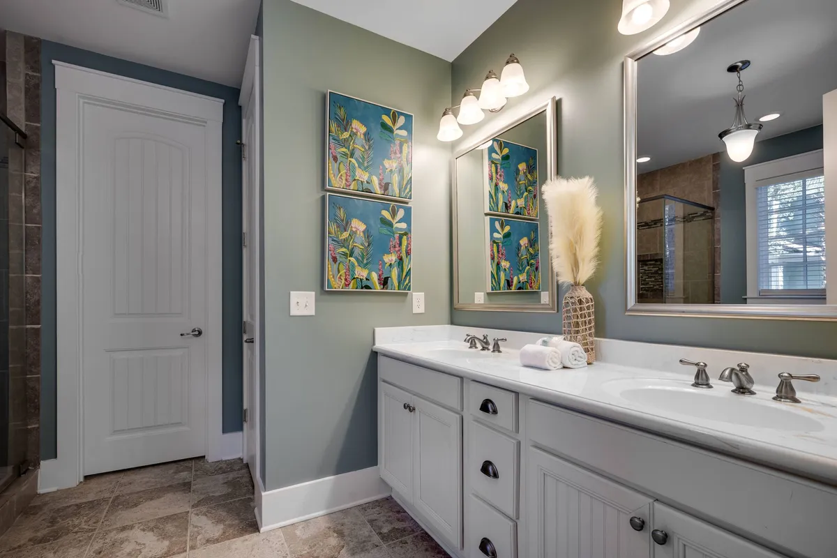 A modern bathroom featuring a vanity with double sinks, large mirrors, elegant lighting, decorative vases, and soft artwork on the walls. The ambiance is bright and serene, providing ample space for morning or evening routines.