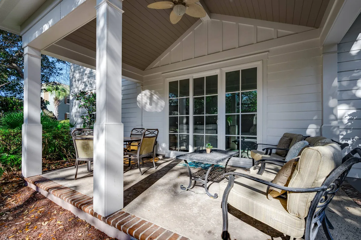 A comfortable and welcoming outdoor seating area is shown, with cushioned chairs arranged around a coffee table. The scene opens to glass doors leading inside, and decorative potted plants add a touch of greenery. The covered patio provides shade and an inviting space for gatherings or enjoying a meal outdoors.