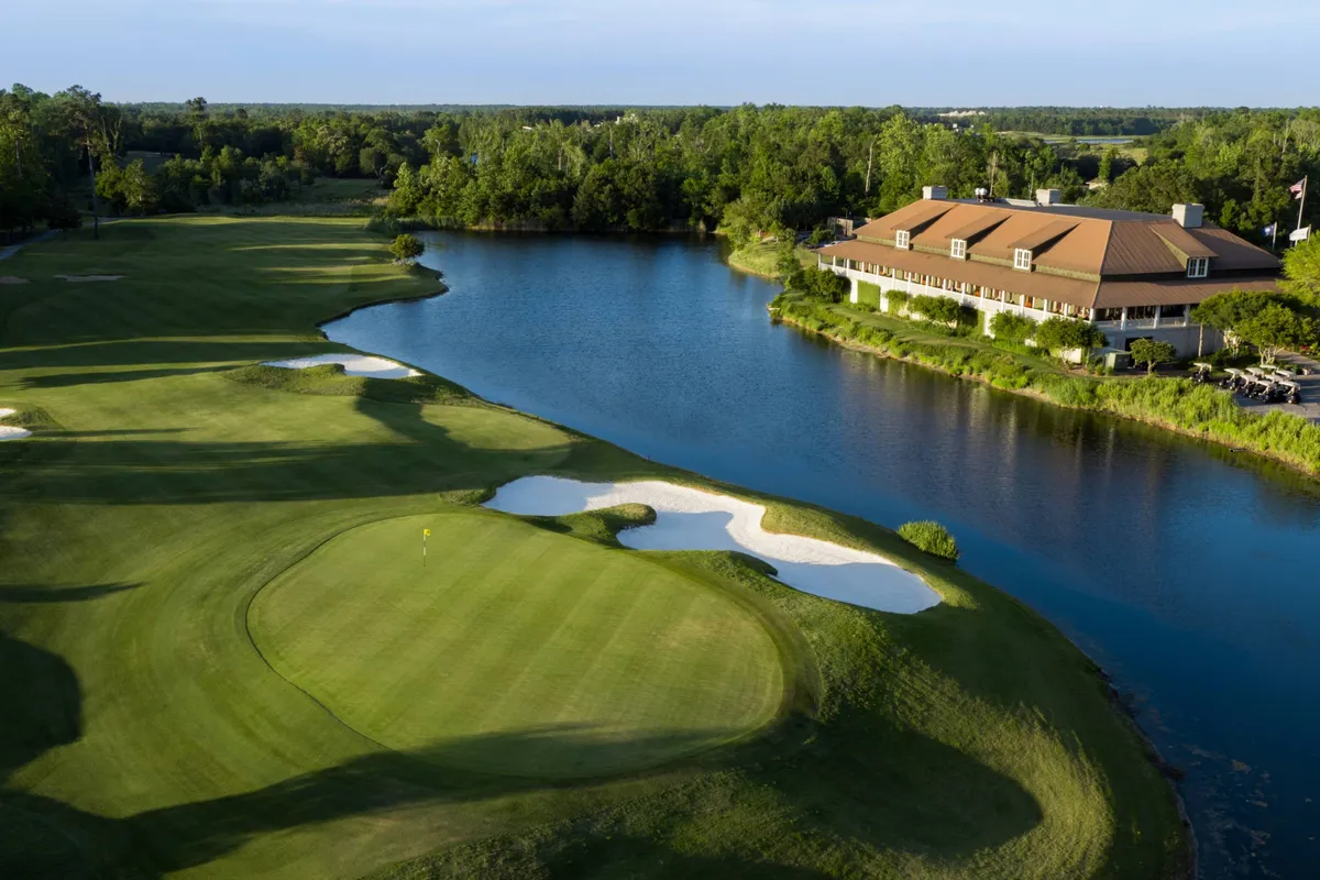 This image captures a lush golf course bordered by a serene lake, with a prominent clubhouse and sand traps visible in the foreground. The scene exudes tranquility, inviting viewers to imagine a day spent golfing in a picturesque setting, socializing at the clubhouse, or simply relaxing by the water after a round.