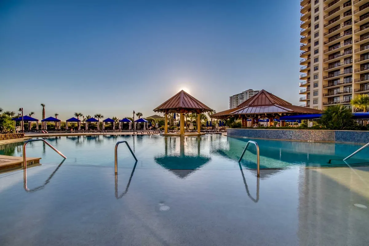 A luxurious pool scene with crystal-clear water, thatched-roof cabanas, and a dramatic sunset in the background. The high-rise resort looms nearby, and guests can be seen enjoying the pool and relaxing by the water, emphasizing both day and evening recreation.