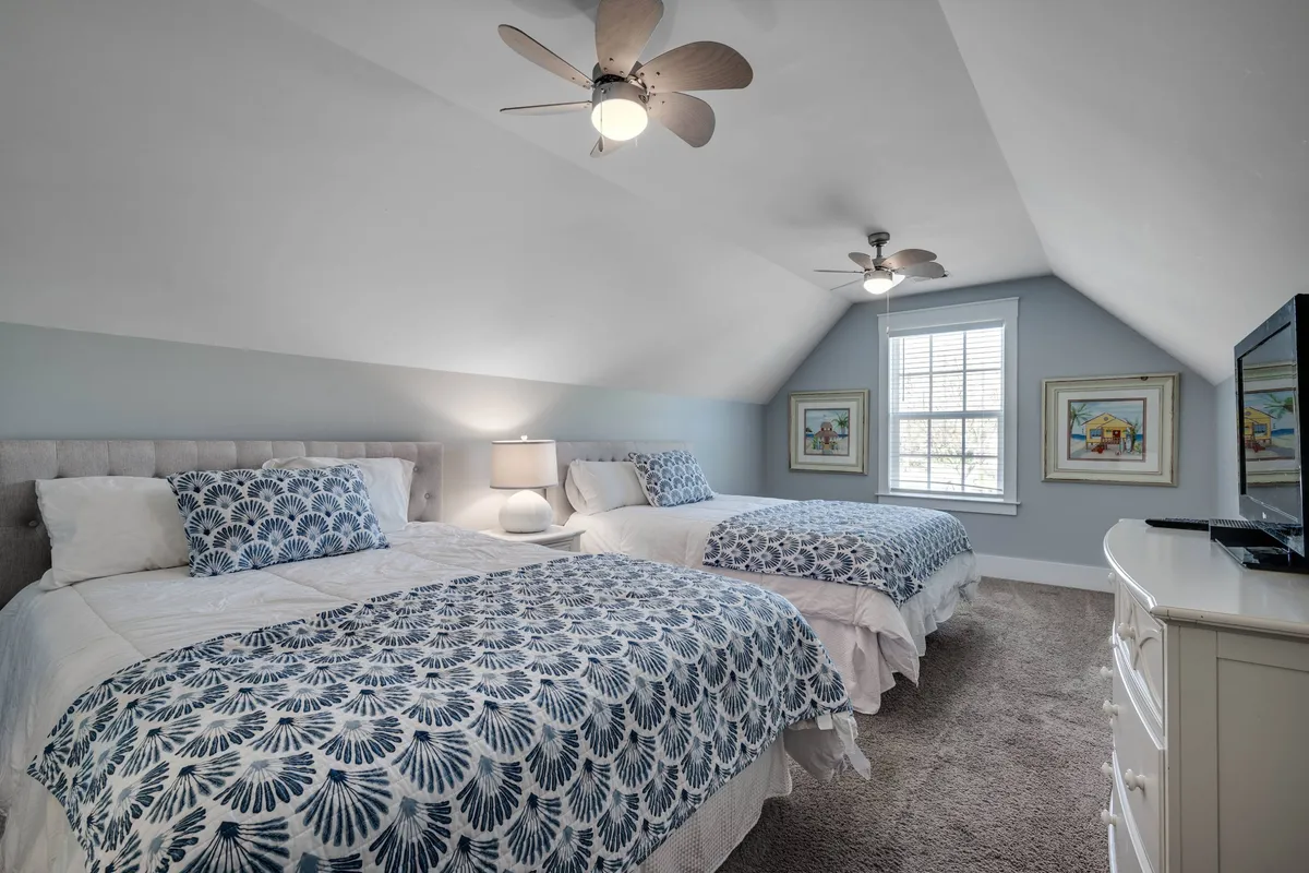 The scene shows a spacious bedroom with two beds dressed in blue and white patterned bedding, a ceiling fan, modern light fixtures, framed artwork, and a television in view. The room is inviting and designed for sharing, making it ideal for families or groups of friends.